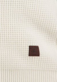 Weißer Pullover aus Waffelstruktur mit geripptem Saum, versehen mit einem braunen Lederetikett "KEY LARGO" mit einem Emblem. Weiches, warmes Material.