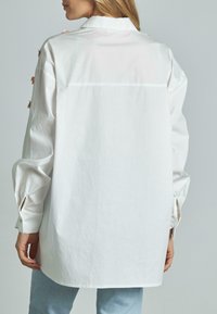 Camicia in cotone bianco con maniche lunghe, colletto a punta, polsini con bottoni e vestibilità rilassata. Presenta ricami floreali sulla spalla.