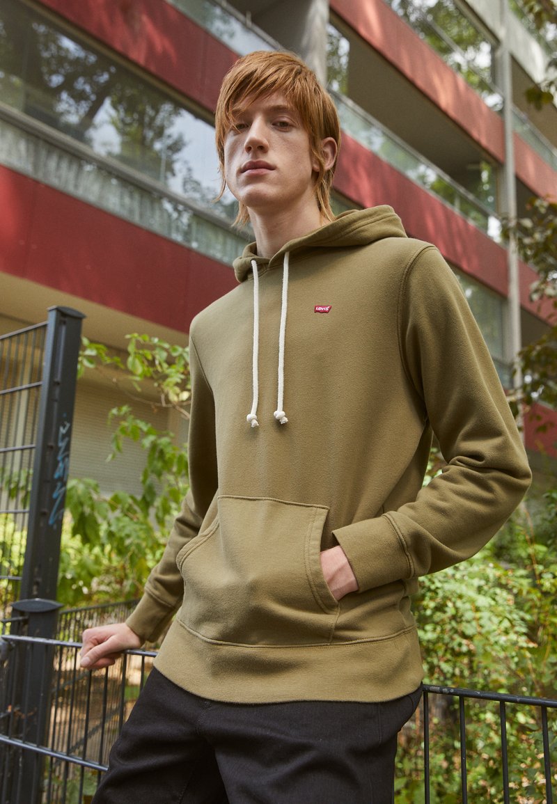 Felpa verde oliva con tasca frontale, cordini bianchi e un piccolo logo rosso Levi's sul petto. Tessuto morbido e testurizzato. Pantaloni neri.