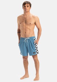 Blaue Badeshorts mit karierten schwarz-weißen Seitenpaneelen, elastischem Bund, Kordelzugverschluss und Logopatch auf der Vorderseite.