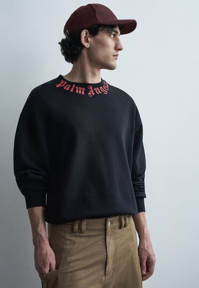 Νεαρός άνδρας που φοράει σκούφο σκούρου κόκκινου χρώματος, μαύρο φούτερ με κόκκινο κείμενο "Palm Angels" και μπεζ παντελόνι, κοιτάζοντας προς τα δεξιά πάνω σε μονόχρωμο φόντο.