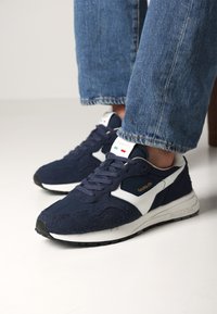 Zapatillas azul marino que combinan ante y tela, con detalles en blanco, cierre de cordones y suela de goma. Detalle del logo en dorado en el lateral.