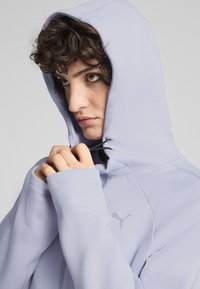 Hoodie violet clair avec capuche à cordon, présentant une texture lisse et un logo discret. Gros plan sur des mains tenant le tissu.