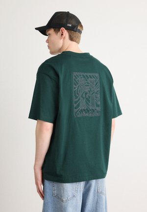 Dypgrønn bomull T-skjorte med korte ermer, med et teksturert svart grafisk design på baksiden, sammen med lyseblå denimshorts.