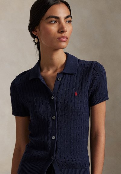 Kobieta w granatowej, krótkim rękawem, rozpinanej koszulce polo z czerwonym logo, patrząca lekko w lewo na jednolitym tle.