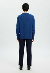 Persona de pie de espaldas, vistiendo un suéter azul, pantalones oscuros y zapatos negros, frente a un fondo claro y liso.