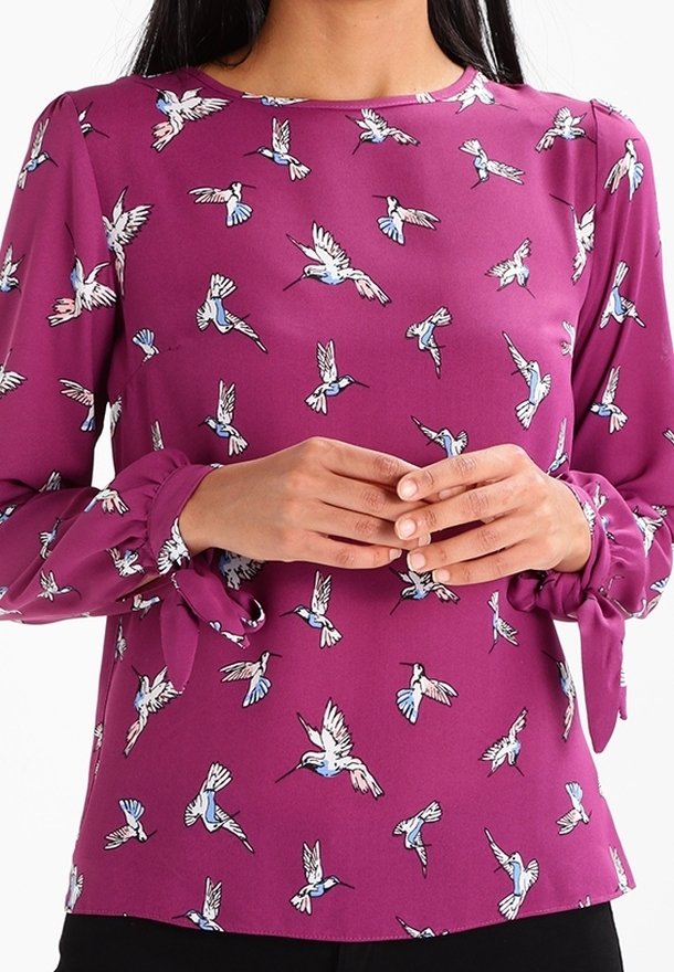 Femme portant un haut à manches longues violet avec des imprimés de colibris bleus et blancs et des poignets noués, mains jointes devant elle.