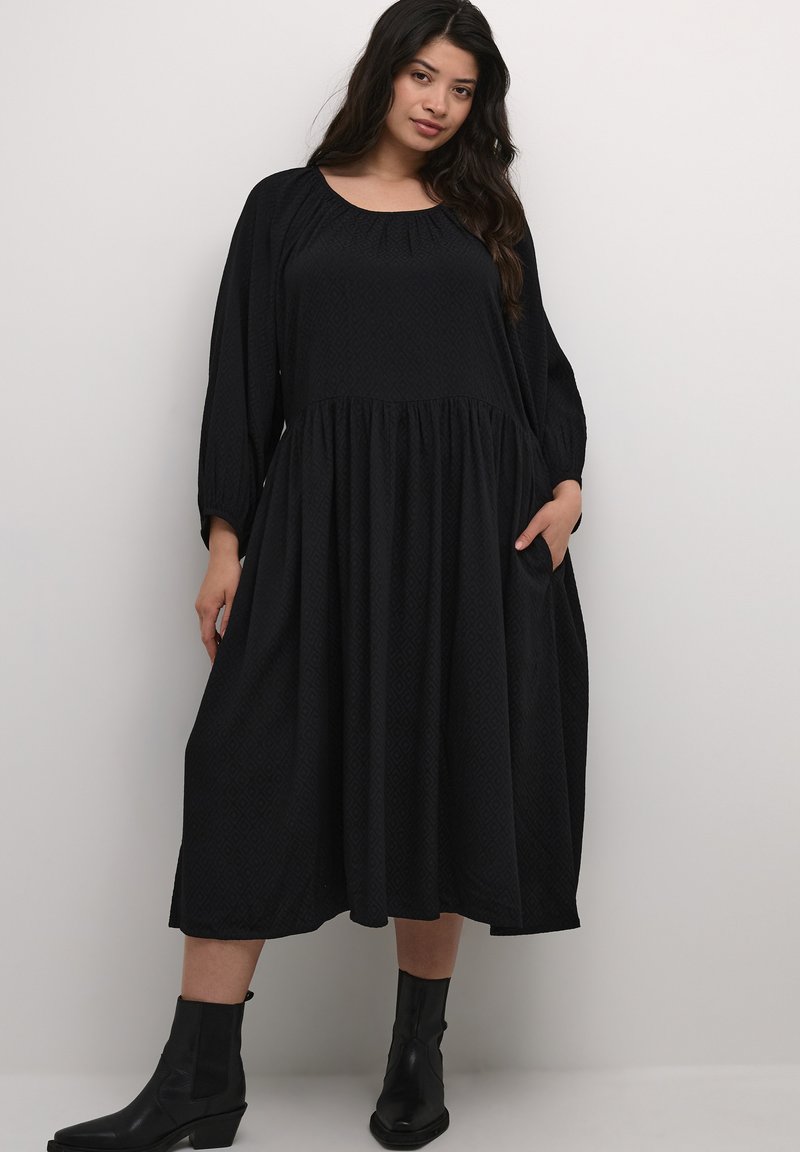 Black midi dress with a scoop neckline, long sleeves, and textured diamond pattern. Features a gathered waist and side pockets. Paired with black ankle boots.