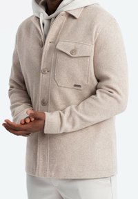 Beige Jacke aus Wollmischung mit Knopfverschluss vorn, zwei Brusttaschen und weicher Textur; darüber getragen ein hellgrauer Hoodie.