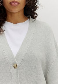 Helle graue Strickjacke mit V-Ausschnitt-Design, ausgestattet mit einem einzelnen runden Knopfverschluss. Gestrickte Textur und lockere Passform über einem weißen T-Shirt.