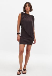 Hnedé, krátke šaty z hladkej látky s prehodeným dizajnom, ktorý má jedno rameno zakryté. V kombinácii s tmavohnedými sandálmi a náramkom.