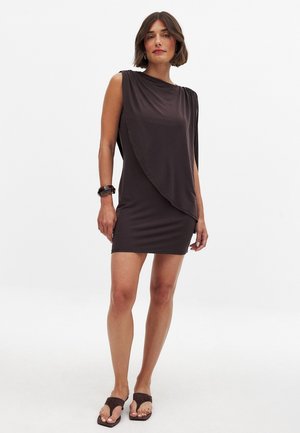 Abito corto marrone realizzato in tessuto morbido, caratterizzato da un design drappeggiato con una spalla coperta. Abbinato a sandali marrone scuro e a un braccialetto.