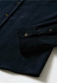 Chemise en velours côtelé bleu marine foncé à manches longues avec des boutons marron, montrant un gros plan du poignet et d'une partie du devant de la chemise sur fond blanc.