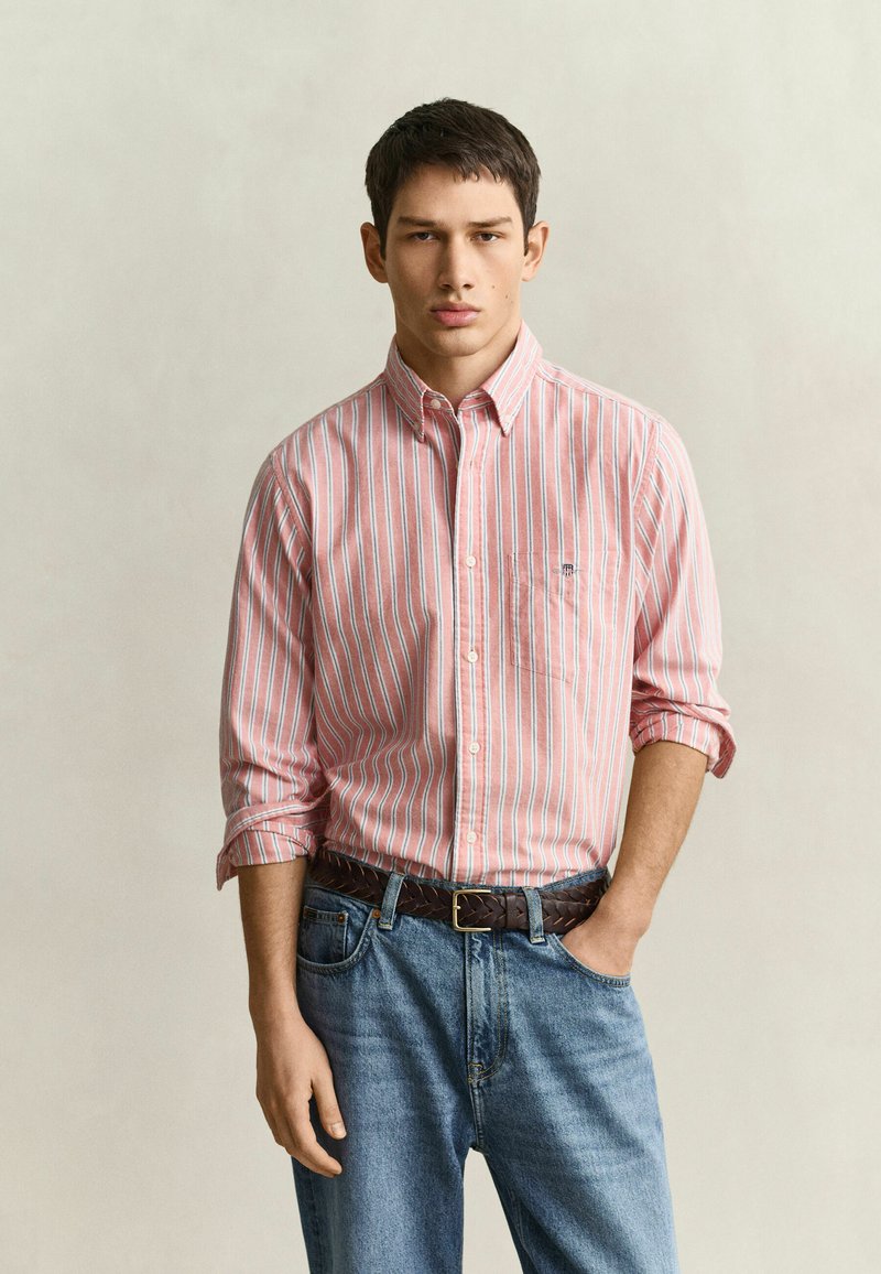 Jeune homme portant une chemise à rayures roses et blanches à manches retroussées, un jean bleu et une ceinture tressée marron, debout devant un fond uni.