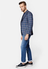 Niebieski blezer w kratę z brązowymi akcentami, w połączeniu z jasnoniebieskimi dżinsami i ciemnymi butami sportowymi. Blezer ma dopasowany krój i klapy typu notch.