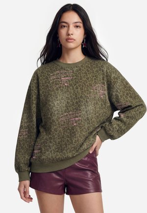 Kvinde med langt mørkt hår iført en grøn leopardmønstret sweatshirt med pink tekst og bordeaux lædershorts, stående med den ene hånd på hoften.