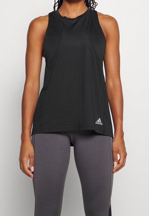 Kobieta ubrana w czarny top Adidas bez rękawów i szare legginsy, stoi ze swobodnie opuszczonymi ramionami wzdłuż ciała.