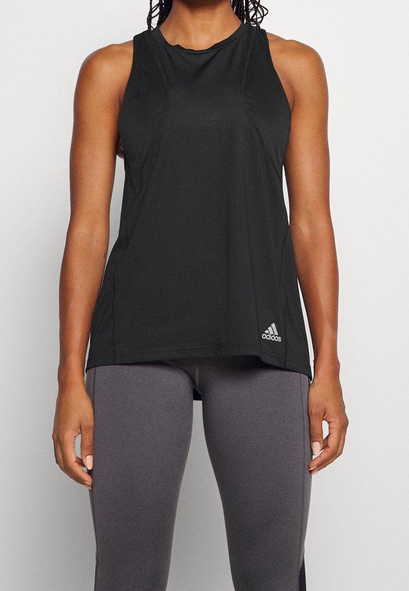 Femme portant un haut Adidas noir sans manches et des leggings gris, debout, les bras détendus le long du corps.