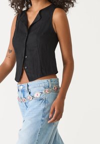 Czarny bezrękawnik z kołnierzykiem, noszony z jasnoniebieskimi dżinsami, do których dodano srebrny i różowy pasek z kwiatowym wzorem.