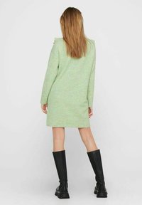 Długa sukienka z dzianiny w zielonym kolorze, o fakturowanym wykończeniu, z prążkowanym dołem i guzikami na ramionach, zestawiona z czarnymi kozakami do kolan.