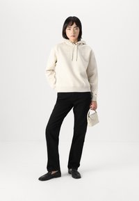 Haut à capuche beige avec un cordon de serrage, présentant un détail de logo subtil. Associé à un pantalon noir et des chaussures noires sans lacets, tenant un sac à main de couleur claire.