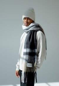 Echarpe en laine à carreaux gris et noirs drapée sur un pull tricoté blanc, accompagnée d'un bonnet blanc. L'étiquette indique "ARCTIC RX." Texture douce.