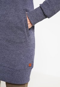 Vestido sudadera azul oscuro con mangas largas, bolsillos laterales, puños acanalados y una tela texturizada. Presenta un pequeño parche de logo de cuero.