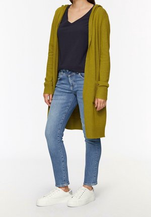 Frau trägt einen olivgrünen, offenen Cardigan, ein schwarzes V-Ausschnitt-Top, hellblaue Skinny-Jeans und weiße Sneakers, steht vor weißem Hintergrund.