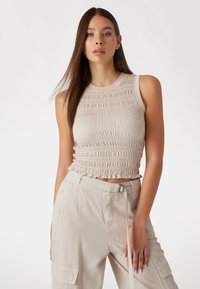 Crop top beige ajusté avec un design texturé et froncé, et un col rond. Porté avec un pantalon cargo beige clair comportant des poches et une ceinture.