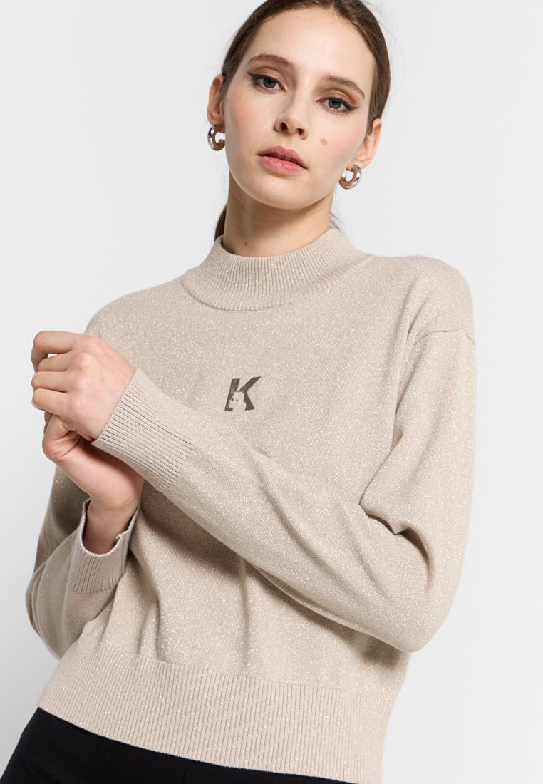 Maglione lavorato a maglia beige con collo alto, polsini e vita a costine. Presenta una sottile texture brillante e un logo "K" marrone sul davanti.