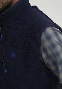 Mornjevo plavi flis prsluk s teksturiranim, dlakavim materijalom i glatkima tkaninama. Ima zip džep, kontrastne šavove i okruglu oznaku sa logotipom.
