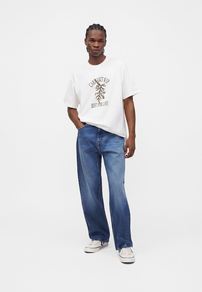 Mann steht mit einer Hand in der Tasche, trägt ein weißes T-Shirt mit Grafik, blaue Jeans und weiße Sneaker vor einem schlichten hellen Hintergrund.