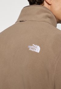 Brown fleece jacket with a high collar. Features a white embroidered logo on the back, smooth texture, and a minimalist design.