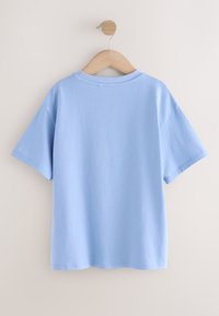 T-shirt di cotone azzurro chiaro a maniche corte appesa a una gruccia di legno contro un muro beige, vista da dietro.