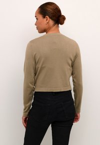 Cárdigan corto de manga larga en punto beige. Presenta un cuello redondo y un dobladillo acanalado, combinado con unos pantalones vaqueros negros de cintura alta. Textura suave.