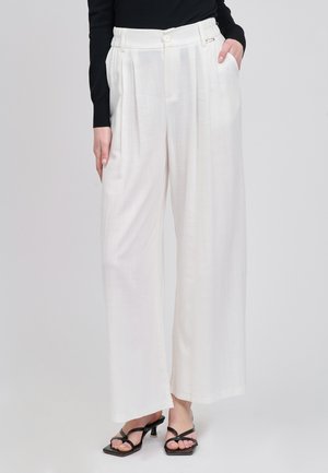 Kvinde iført hvide bukser med højtaljet vidde og plisseringer, sidelommer, kombineret med sorte hælede sandaler med stropper og en sort top.