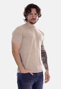 Maglione beige a maniche corte con scollatura rotonda, tessuto testurizzato e vestibilità aderente, abbinato a jeans di denim scuri, indossato da un uomo.