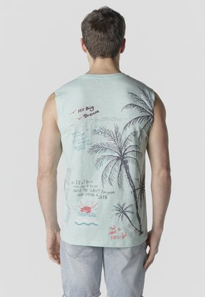 Hombre vistiendo una camiseta sin mangas azul claro con palmeras y texto de temática tropical visto desde atrás, combinado con pantalones cortos de mezclilla claros.