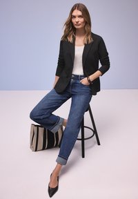 Femme assise sur un tabouret portant un blazer noir, un haut blanc, un jean bleu retroussé, des talons noirs, la main dans la poche, avec un sac rayé posé au sol.