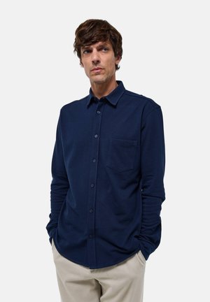 Chemise à manches longues marine en tissu doux, design à boutons, poche avant unique, col classique, coupe ajustée, portée avec un pantalon beige clair.