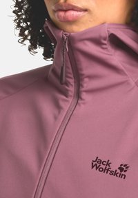 Giacca in pile di un morbido color malva con colletto alto, logo nero e chiusura lampo a vista con cursore. Texture liscia e design aderente.