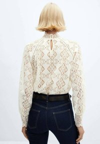 Blonde bluse i creme med geometriske mønstre, lange ærmer og en nøglehul-rygdetalje, parret med mørk denim jeans og et sort bælte.