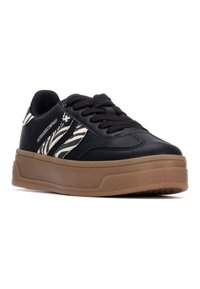 Zapatilla de cuero negra con rayas zebra blancas, punta redonda, suela gruesa de goma marrón y cordones negros. El texto incluye el logo de la marca en el costado.