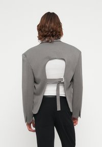Gray oversized blazer with a cut-out back and adjustable strap detail. Styled over a white top, paired with black pants. Smooth texture.
