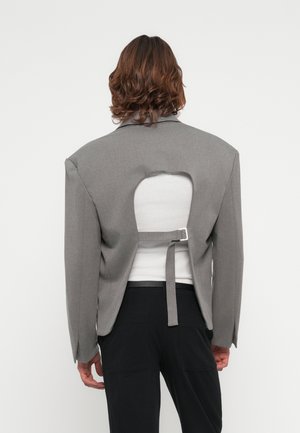 Gray oversized blazer with a cut-out back and adjustable strap detail. Styled over a white top, paired with black pants. Smooth texture.