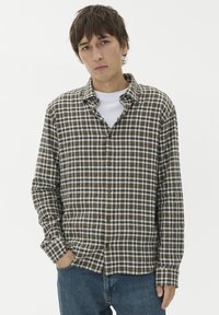 Brązowa i biała koszula w kratę z długimi rękawami, z kołnierzykiem, z brązowymi guzikami z przodu. Nosi się ją na białej koszulce z okrągłym dekoltem.