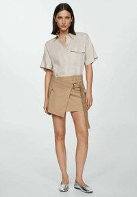 Beige shirt with short sleeves and a chest pocket, paired with a tan wrap skirt featuring a buckle and buttons. Silver flat shoes.