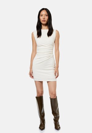 Kvinde med langt mørkt hår iført en hvid ærmeløs minikjole og mørkebrune læderstøvler op til knæet, stående mod en ensfarvet baggrund.