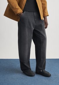 Άνδρας που φοράει φαρδιά μαύρη παντελόνια με μεγάλες μπροστινές τσέπες, μαύρα παπούτσια, μαύρο πουκάμισο και καφέ σακάκι, στέκεται σε μπλε χαλί.