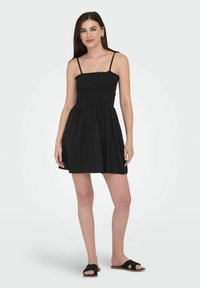 Robe noire avec un corsage smocké, des bretelles fines et une jupe évasée. Portée avec des sandales plates noires. Tissu doux, détails minimalistes.